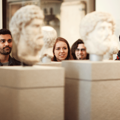 Visitors to the museum exploring sculpture in the gallery. Their faces can be seen between stone heads.